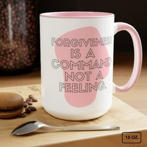 May include: White ceramic mug with a pink rim and handle. The mug features a pink abstract shape with the text "FORGIVENESS IS A COMMAND NOT A FEELING." in black lettering. The mug is 15 ounces.