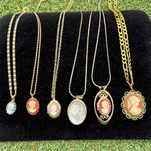 May include: A collection of gold-toned necklaces with cameo pendants. The pendants feature carved portraits in various colors, including blue, orange, and white. The necklaces are displayed on a black surface against a backdrop of green grass.