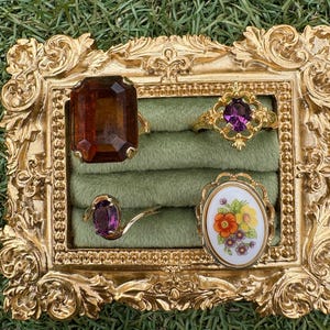 Puede incluir: Una colección de cuatro anillos exhibidos en un marco dorado y ornamentado. Los anillos presentan varios diseños, incluyendo una gran gema marrón, una gema púrpura y un diseño floral. El marco se encuentra sobre una base de tela verde.