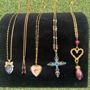 May include: A collection of gold-toned necklaces with various pendants, including a heart-shaped crystal, a purple gemstone, a heart locket with a rose, and a cross with blue gemstones. The necklaces are displayed on a black surface.