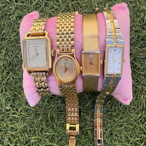 May include: Four vintage wristwatches displayed on a pink cushion. The watches feature gold and silver tones, with rectangular and round faces. One watch has a square face and a gold strap. The watches are set against a backdrop of green grass.