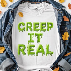 May include: White t-shirt featuring the phrase "CREEP IT REAL" in dripping green lettering. The shirt is set against a denim jacket background with autumnal leaves, giving a seasonal, spooky feel.