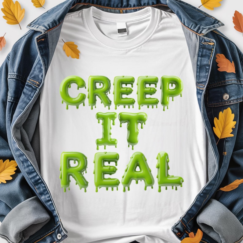 May include: White t-shirt featuring the phrase "CREEP IT REAL" in dripping green lettering. The shirt is set against a denim jacket background with autumnal leaves, giving a seasonal, spooky feel.