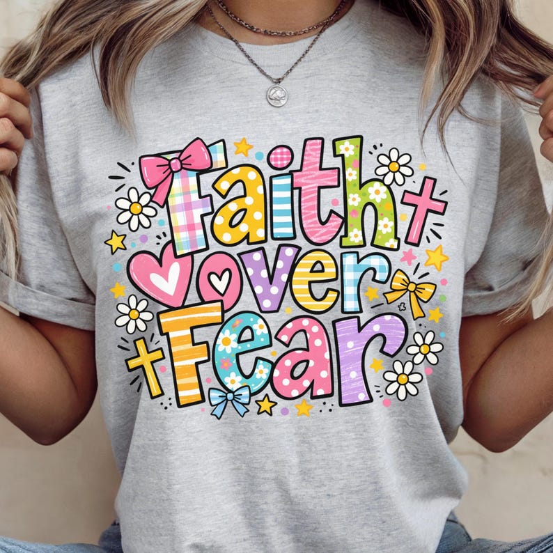 May include: A light gray t-shirt featuring the phrase "Faith Over Fear" in a colorful, whimsical design. The text is surrounded by daisies, stars, bows, and hearts. The shirt has short sleeves and a crew neck.