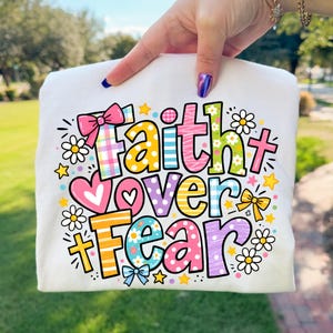 May include: White t-shirt with the words "Faith Over Fear" in colorful, playful lettering. The design includes hearts, stars, bows, and crosses. The shirt is held up by a person.