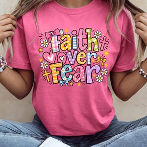 May include: A pink t-shirt with the words "Faith Over Fear" in a colorful, whimsical design. The text includes hearts, bows, stars, and flowers. The shirt is short-sleeved.