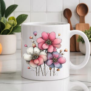 Puede incluir: Taza de cer&aacute;mica blanca con un dise&ntilde;o floral en acuarela. El dise&ntilde;o incluye tres flores grandes en tonos rosa, blanco y azul, con delicados tallos y capullos. La taza tiene un asa est&aacute;ndar y est&aacute; sobre un fondo neutro.
