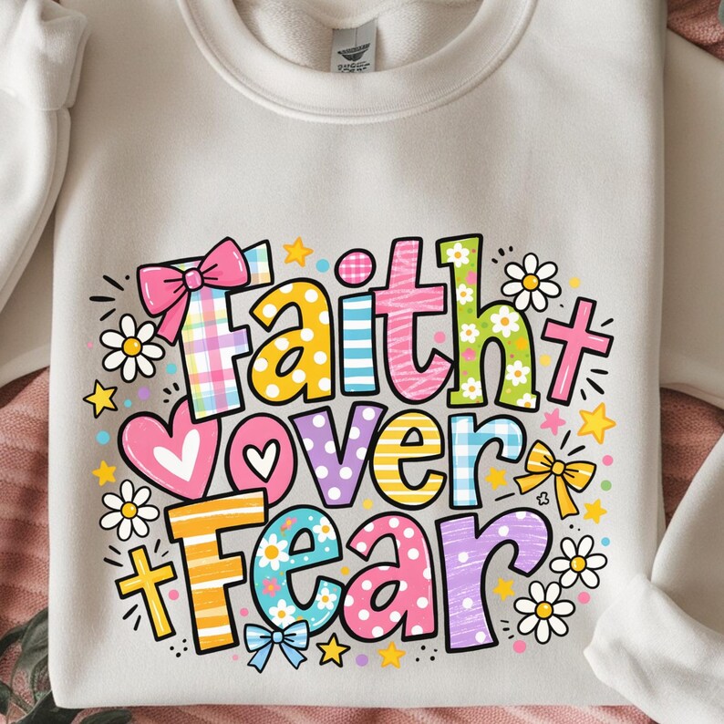 May include: Cream-colored sweatshirt with a colorful graphic that reads "Faith Over Fear." The design includes hearts, stars, bows, and flowers. The text is in various colors and patterns, including plaid and floral.