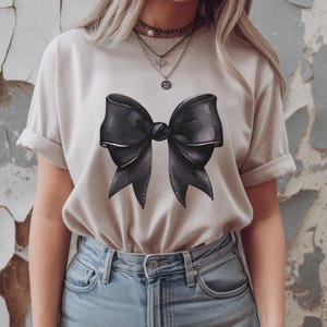 May include: A light beige t-shirt with a large black bow graphic printed on the front.