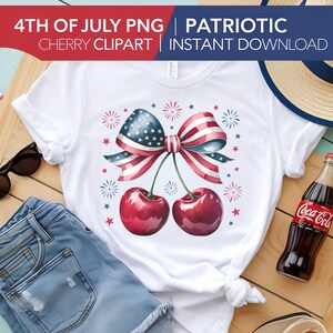 May include: A white t-shirt with a red, white, and blue cherry design. The cherries are tied together with a bow made of an American flag. The design is surrounded by red, white, and blue stars and fireworks.