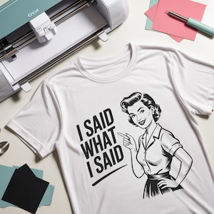 May include: White t-shirt with the text "I SAID WHAT I SAID" and a black and white illustration of a woman pointing. A Cricut machine and craft supplies are in the background.