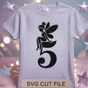 May include: A light purple t-shirt featuring a black silhouette of a fairy sitting on the number 5. The text "SVG CUT FILE" is displayed at the bottom of the shirt. The shirt has short sleeves and a crew neck.
