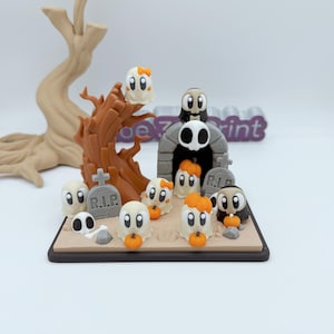 May include: A Halloween-themed diorama featuring cartoon ghosts, pumpkins, tombstones, and a spooky archway. The ghosts have various accessories, including bows and capes. The scene is set on a rectangular base with a brown tree and a light-colored background.