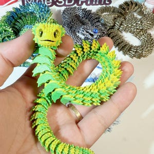 May include: A green and yellow 3D printed origami dragon with a smiling face. The dragon is posed in a curled position with its tail curled around its body. The dragon is made of many small, overlapping scales.