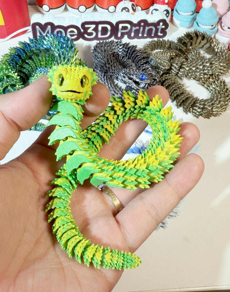 May include: A green and yellow 3D printed origami dragon with a smiling face. The dragon is posed in a curled position with its tail curled around its body. The dragon is made of many small, overlapping scales.
