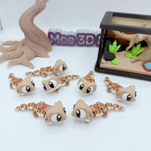 Pu&ograve; includere: Una collezione di figurine di geco marroni e beige con grandi occhi espressivi. Le figurine sono disposte su una superficie bianca, con un terrario in miniatura e un albero decorativo sullo sfondo. Il testo "Moe3D" &egrave; visibile.