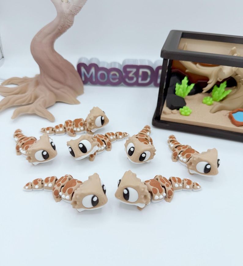 Pu&ograve; includere: Una collezione di figurine di geco marroni e beige con grandi occhi espressivi. Le figurine sono disposte su una superficie bianca, con un terrario in miniatura e un albero decorativo sullo sfondo. Il testo "Moe3D" &egrave; visibile.