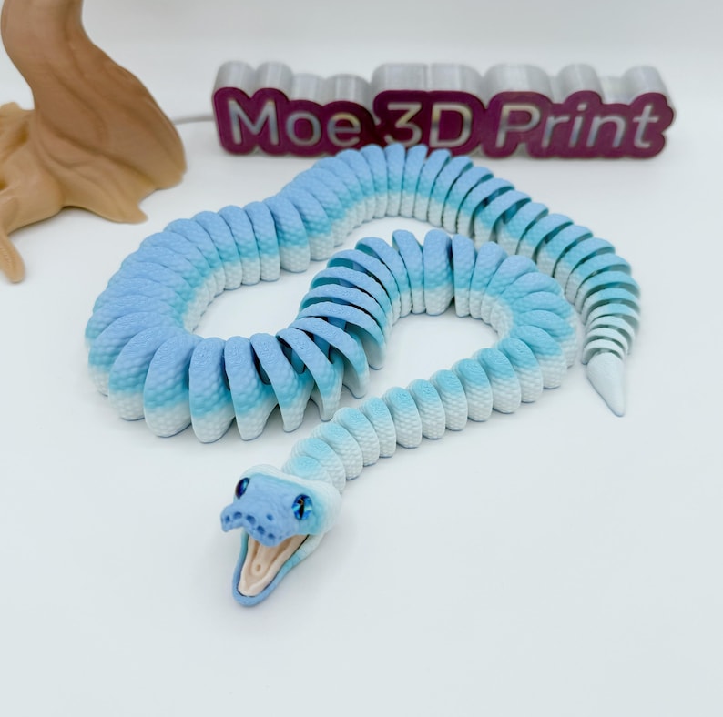 Op de afbeelding: Een 3D-geprint gearticuleerd slangen speelgoed in tinten blauw en wit. De slang heeft een gedetailleerde kop met blauwe ogen en een open mond. De tekst "Moe 3D Print" is zichtbaar op de achtergrond.