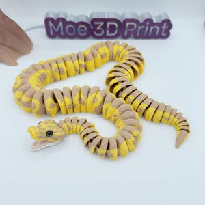 Op de afbeelding: Een 3D-geprint gearticuleerd slangen speelgoed in geel en bruin. Het lichaam van de slang is gesegmenteerd, waardoor flexibele beweging mogelijk is. De kop heeft gedetailleerde ogen en een licht geopende mond. De tekst "Moe 3D Print" is zichtbaar op de achtergrond.