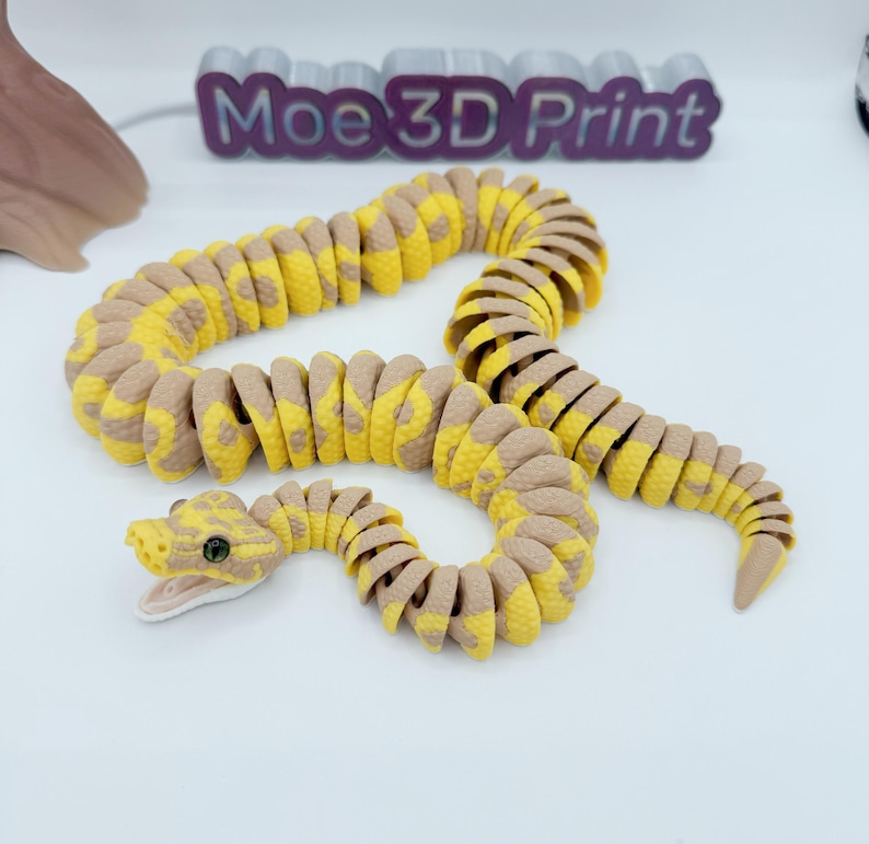 Op de afbeelding: Een 3D-geprint gearticuleerd slangen speelgoed in geel en bruin. Het lichaam van de slang is gesegmenteerd, waardoor flexibele beweging mogelijk is. De kop heeft gedetailleerde ogen en een licht geopende mond. De tekst "Moe 3D Print" is zichtbaar op de achtergrond.