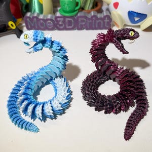 May include: Two 3D printed snake figurines, one blue and white, the other purple and black.  The snakes are curled into a circle shape.  The text "Moe 3D Print" is visible in the background.
