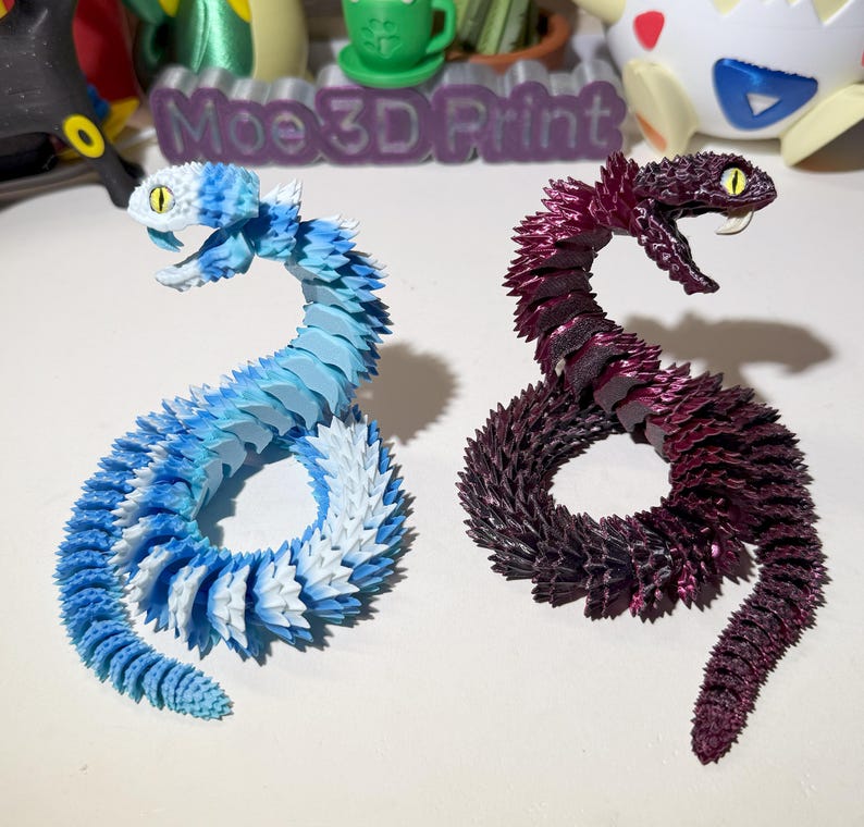 May include: Two 3D printed snake figurines, one blue and white, the other purple and black.  The snakes are curled into a circle shape.  The text "Moe 3D Print" is visible in the background.