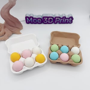 May include: Two egg cartons, one white and one brown, each holding six colorful eggs. The white carton contains yellow, pink, and white eggs. The brown carton holds green, blue, and pink eggs. The text "Moe 3D Print" is visible in the background.
