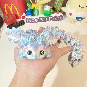 May include: A light blue and iridescent 3D printed dragon figurine with a long tail and a cute face. The dragon has a textured, scaly body and is holding a flower in its mouth. The dragon is sitting on a hand.