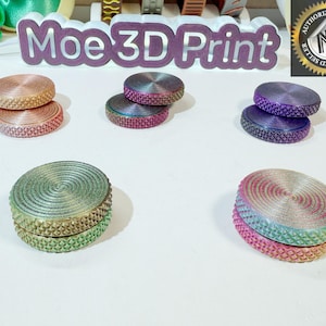 May include: Six different color combinations of a two-piece fidget spinner. The spinners are made of a smooth, iridescent plastic with a textured outer ring. The spinners are stacked on top of each other, with the top piece slightly offset from the bottom piece. The spinners are arranged in a circle on a white surface. The text "Moe 3D Print" is visible in the background.