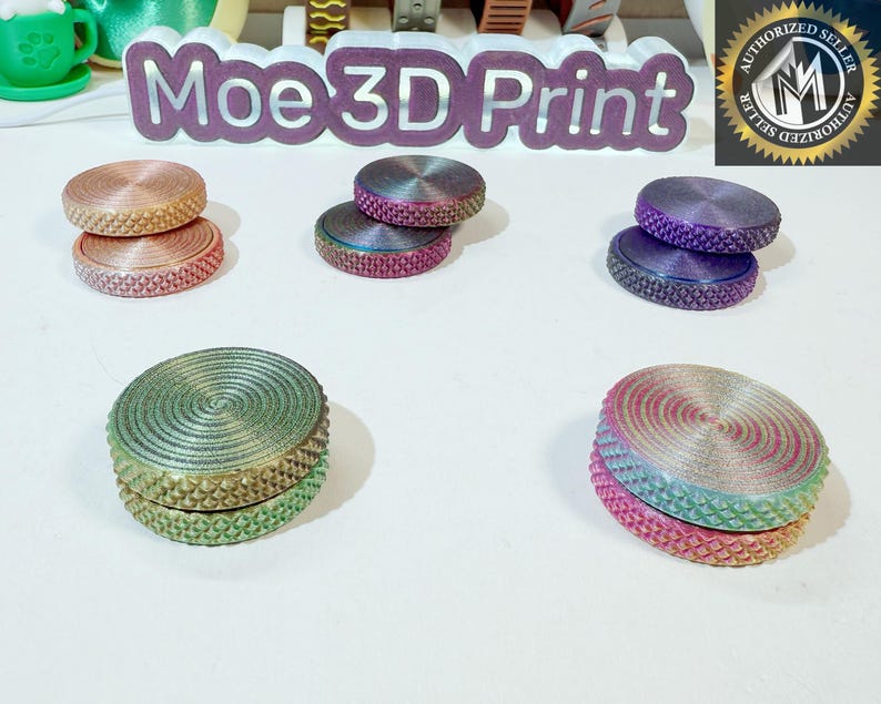 May include: Six different color combinations of a two-piece fidget spinner. The spinners are made of a smooth, iridescent plastic with a textured outer ring. The spinners are stacked on top of each other, with the top piece slightly offset from the bottom piece. The spinners are arranged in a circle on a white surface. The text "Moe 3D Print" is visible in the background.