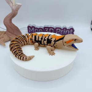 May include: A detailed 3D-printed lizard figurine with a tan head and body, and a segmented tail. The lizard features orange and black markings along its back and tail, with a blue mouth. The base is white, and the text "Moe 3D Print" is visible in the background.