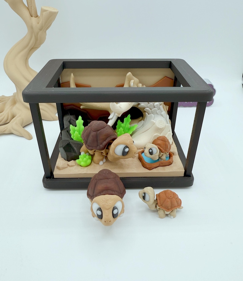 Pu&ograve; includere: Un diorama con una famiglia di tartarughe dei cartoni animati. La scena comprende una scatola con cornice nera con una base sabbiosa, piante verdi e un tronco di colore chiaro. Diverse tartarughe sono all'interno della scatola e due sono all'esterno. Le tartarughe hanno gusci marroni e grandi occhi espressivi.