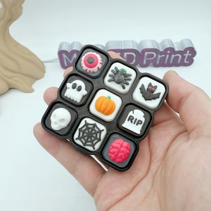 May include: A black Halloween-themed tray with nine square compartments, each containing a different spooky icon. Icons include a white skull, a spider, a pumpkin, a bat, and a tombstone. The text "M 3D Print" is visible in the background.