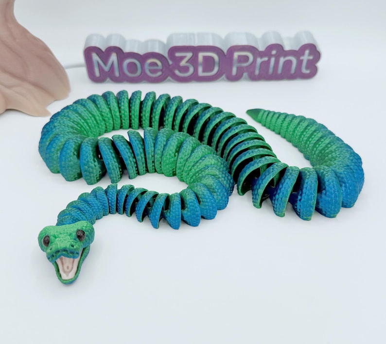 Op de afbeelding: Een 3D-geprint, beweeglijk slangen speelgoed in blauw- en groentinten. De slangenkop is groen met een open mond, en het lichaam gaat over van groen naar blauw. De tekst "Moe 3D Print" is zichtbaar op de achtergrond.