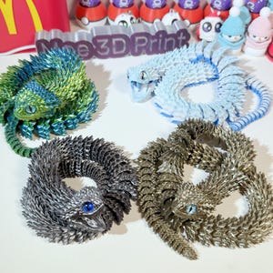 May include: Four 3D printed lizard figurines in different colors: green, blue, silver, and gold. Each lizard is curled up in a circle with its tail in its mouth.