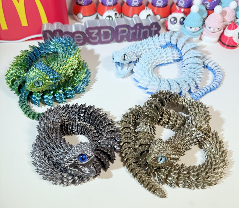 May include: Four 3D printed lizard figurines in different colors: green, blue, silver, and gold. Each lizard is curled up in a circle with its tail in its mouth.