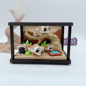 以下が含まれることがあります： 3匹の漫画のヤモリが登場するミニチュアテラリウムのシーン。 ヤモリは茶色と茶色の斑点で、木の枝、砂、緑の植物を背景にしています。 テラリウムは濃い茶色のフレームに囲まれています。