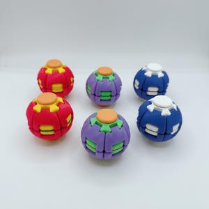 May include: A collection of five spherical fidget toys arranged in two rows. Two are red with yellow accents, two are purple with green accents, and one is blue with white accents. Each sphere features a central button on the top surface.