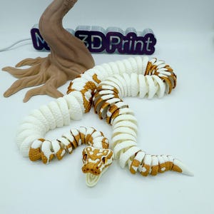 Op de afbeelding: Een 3D-geprint gearticuleerd slangen speelgoed in wit en bruin. De slang is gesegmenteerd, waardoor flexibele beweging mogelijk is. De kop heeft gedetailleerde ogen en een realistisch ontwerp. De achtergrond bevat een boomsculptuur en de tekst "3D Print".