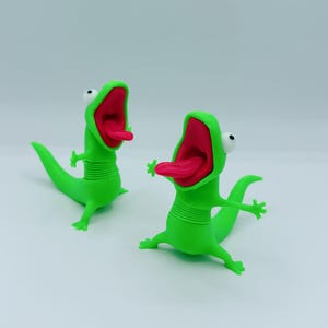 May include: Two bright green lizard figurines with wide-open mouths, protruding pink tongues, and large white eyes with black pupils. Each lizard features a flexible, ribbed midsection, four small limbs, and a long tail, standing in an upright pose.