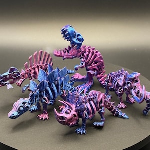 Puede incluir: Cinco esqueletos de dinosaurios impresos en 3D en varias poses. Los esqueletos están pintados en un degradado de morado y azul.