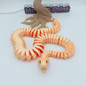 Op de afbeelding: Een 3D-geprint, beweeglijk slangen speelgoed in perzik- en oranje tinten, met een gedetailleerde kop en gele ogen. De slang is opgerold, met een boomachtige structuur en de tekst "Mo Print" op de achtergrond.