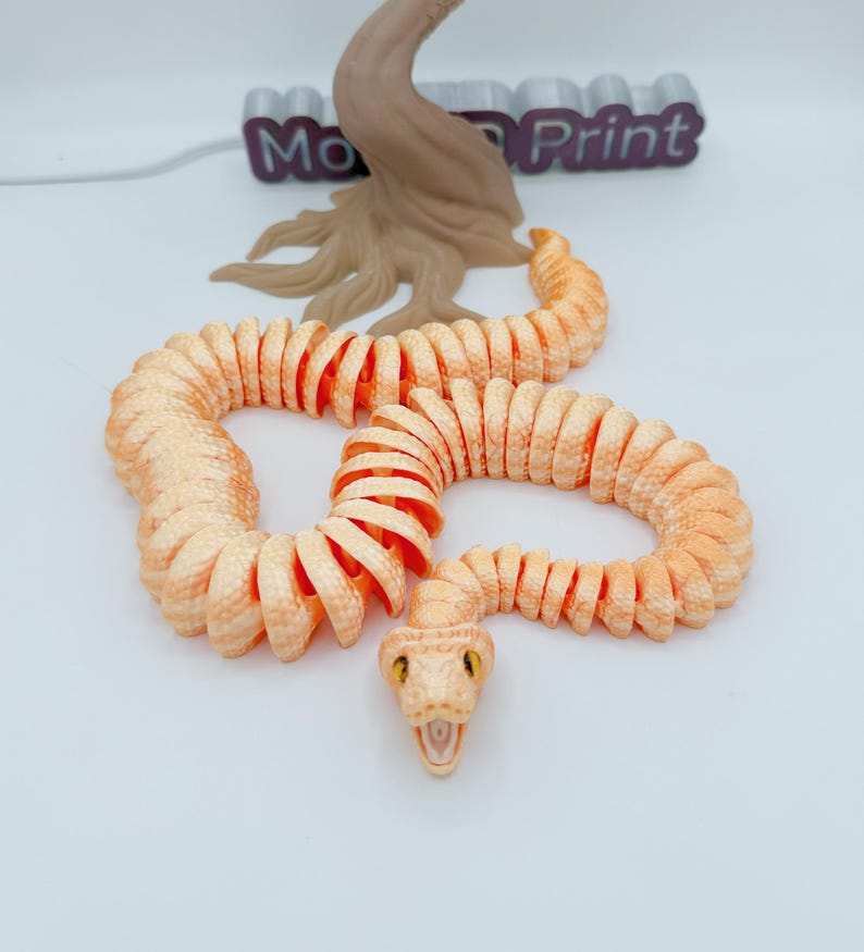 Op de afbeelding: Een 3D-geprint, beweeglijk slangen speelgoed in perzik- en oranje tinten, met een gedetailleerde kop en gele ogen. De slang is opgerold, met een boomachtige structuur en de tekst "Mo Print" op de achtergrond.