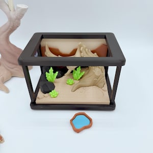 Pu&ograve; includere: Un terrario in miniatura con una cornice nera, con un paesaggio desertico con sabbia beige, formazioni rocciose marroni e piante verdi. Un piccolo stagno blu con un bordo marrone &egrave; di fronte.