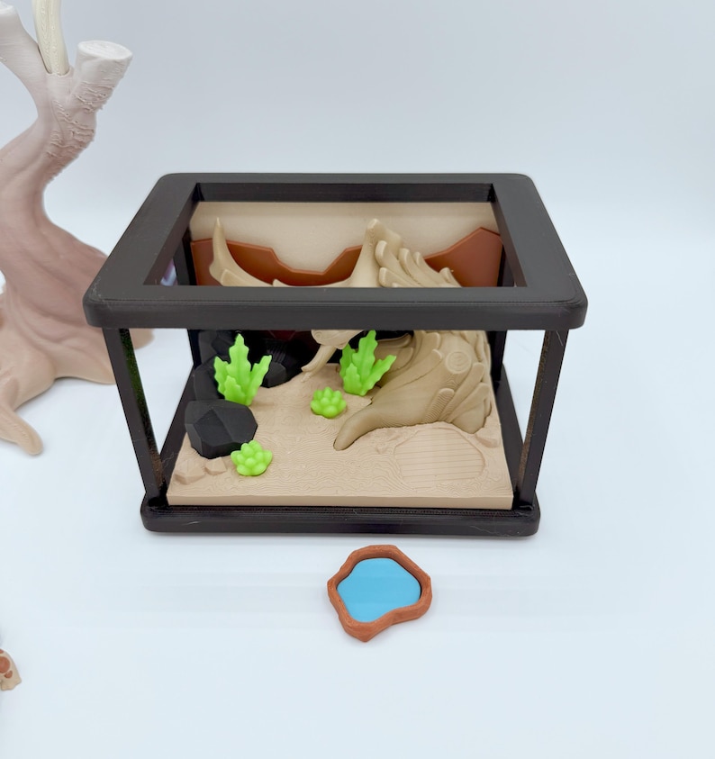Pu&ograve; includere: Un terrario in miniatura con una cornice nera, con un paesaggio desertico con sabbia beige, formazioni rocciose marroni e piante verdi. Un piccolo stagno blu con un bordo marrone &egrave; di fronte.