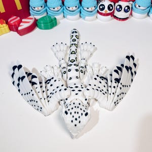 May include: A white and black spotted, articulated, 3D printed dragon with yellow and green markings on its tail. The dragon has wings that are folded back and is lying on its back.