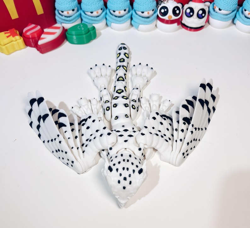 May include: A white and black spotted, articulated, 3D printed dragon with yellow and green markings on its tail. The dragon has wings that are folded back and is lying on its back.