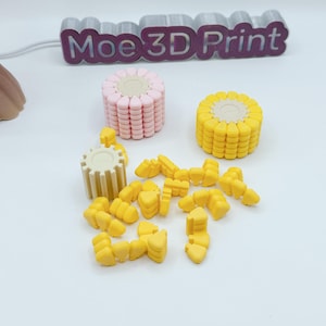 Puede incluir: Un conjunto de objetos impresos en 3D con forma de maíz. Incluye una mazorca rosa y otra amarilla, granos amarillos individuales y una pieza blanca en forma de engranaje. El texto "Moe 3D Print" es visible.