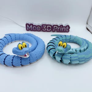 Puede incluir: Dos serpientes de juguete impresas en 3D en tonos azules, enrolladas sobre una superficie blanca. Una es azul sólido, la otra es una mezcla de azul y verde. Ambas tienen ojos amarillos, rayas negras y bocas abiertas. El texto "Moe 3D Print" es visible.