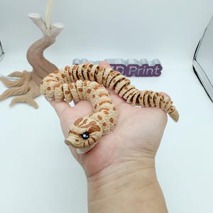 May include: A tan and brown articulated snake toy with a detailed head and body. The snake is held in a hand, showcasing its flexible design. The snake's body is segmented with brown spots.